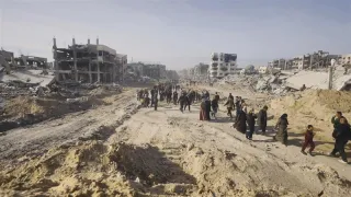 Pessoas deslocadas caminham em terreno desértico, com prédios destruídos ao fundo.