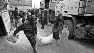Two women carry a large sack of aid supplies together in front of Red Cross trucks, while other people unload and distribute relief items in the background