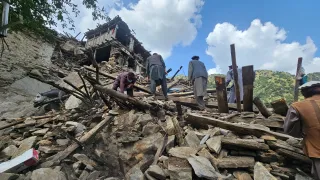 People search through the rubble of collapsed buildings after an earthquake, surrounded by broken wooden beams and stones, with mountains visible in the background under a partly cloudy sky.