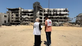 Two humanitarian workers wearing vests with Red Cross and partner organization logos stand in front of a heavily damaged, partially destroyed building, assessing the destruction in Gaza