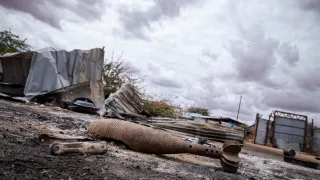 A rusted, unexploded mortar round lies on scorched ground in an abandoned area of Gedo’s Luuq district, Somalia. Behind it, twisted metal sheets and damaged structures sit under a heavy, overcast sky, highlighting the destruction left by recent fighting.