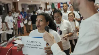 A woman speaks into a microphone while holding a paper that reads “ARMENIA #NotAloneInSiles #WeStandWithYou,” during an indoor group event. Several people in white shirts stand behind her, some holding papers, with international flags visible in the background.
