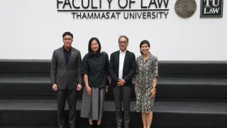 Four people standing and posing for a group photo in front of a wall that reads “Faculty of Law, Thammasat University,” with the university’s emblems displayed above them.