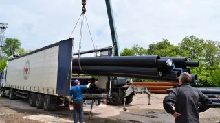 Workers use a crane to unload long black pipes from a truck at an outdoor work site, with one person guiding the load and others observing nearby.