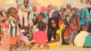 A group of displaced families sits together on sandy ground in Tawila, Sudan. Several women and children gather closely, some looking toward the camera. In the foreground, a young boy stands holding a piece of paper while another child in a headscarf sits among the group. The scene reflects the difficult conditions people are facing after fleeing violence.