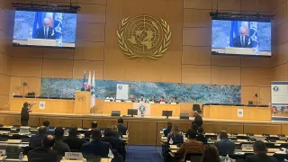 A large conference hall with delegates seated at long rows of desks facing a stage. Two large screens display a speaker at a podium, with the United Nations emblem prominently mounted on the wall behind him. Several panelists sit at a long table on the stage beneath a wide mural. Flags and conference banners are positioned to the side. The audience listens as a photographer captures the scene.