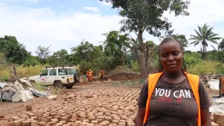Una mujer mira a la cámara. A sus espaldas hay ladrillos, más personas de la comunidad y un carrod del CICR.