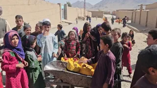 A group of children gather around a wheelbarrow carrying yellow jerrycans in a dusty street, with buildings and mountains in the background.