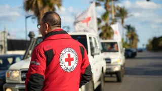 An ICRC staff member wearing a red jacket with the Red Cross emblem stands beside a line of Red Cross vehicles on a coastal road in Lebanon during humanitarian operations amid escalating hostilities.