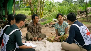 ICRC staff and volunteers sit with two local community members on a mat outdoors in a rural setting