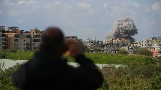 Una persona está de pie en primer plano, desenfocada y vista desde atrás. Sosteniendo un teléfono mientras mira hacia una ciudad donde una gran columna de humo se eleva sobre los edificios a lo lejos.