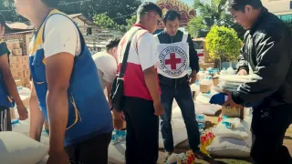 Aid workers distribute relief supplies to people gathered outdoors, with a Red Cross volunteer assisting at a table stacked with bags of goods.