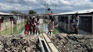 Personas, entre ellas varios niños, se encuentran en un asentamiento con viviendas precarias, donde el agua estancada ha inundado el área. Un estrecho paso improvisado con tablas cruza sobre el agua, mientras las casas de ambos lados muestran condiciones deterioradas bajo un cielo parcialmente nublado.