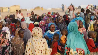 Crowd of women and children gathered outdoors in Tawila, North Darfur, many seated on the ground wearing colorful headscarves and traditional clothing, with a few people standing in the background against a dry, sandy landscape.