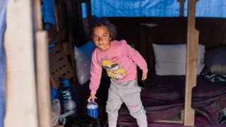 A young girl stands inside a makeshift shelter, holding a small blue container, with bedding and personal belongings arranged around her.