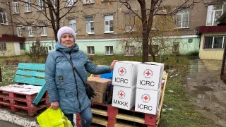 Woman in a blue coat and knit hat stands outdoors beside a stack of white aid boxes marked with red cross symbols on wooden pallets, holding a bright yellow bag; apartment buildings and bare trees are in the background.
