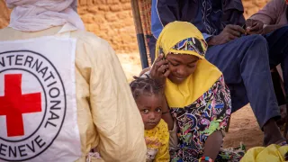 A woman wearing a yellow headscarf sits and speaks on a phone, with a young child beside an ICRC vest in Anderamboukane, Mali, as people gather in the background outdoors.