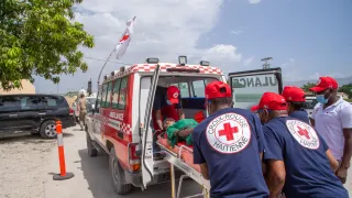 Personal de la Cruz Roja Haitiana traslada a una persona herida en camilla hacia una ambulancia. Varios socorristas participan en la evacuación y atención de emergencia durante el traslado del paciente.