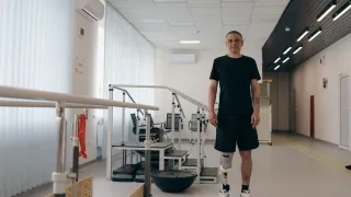 A man with two lower-leg prosthetic limbs stands in a rehabilitation centre beside parallel bars used for gait training. Exercise equipment is visible in the background of the bright therapy room.
