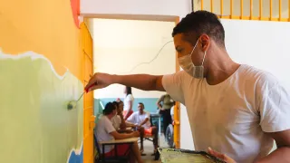 Una persona privada de libertad, con mascarilla, pinta una pared con un rodillo dentro de un centro de detención. En el fondo, otras personas participan en la actividad mientras permanecen en un pasillo del establecimiento.