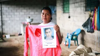 A woman holds a red shirt and a missing person poster featuring a photograph of Rigoberto Lipe Ocotán, who has been missing since June 13, 2020. She looks directly at the camera from inside her home.