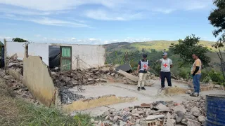 Severely damaged home with collapsed walls and rubble on the ground. Two International Committee of the Red Cross staff members speak with a woman while assessing the damage. A mountainous landscape can be seen in the background.