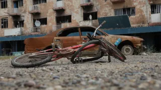 A damaged bicycle lies on a gravel-covered street in front of a burnt-out car and a fire-scorched apartment building, showing destruction in an urban neighbourhood after fighting.