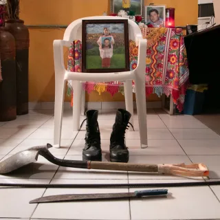 Una foto de una mujer sosteniendo un retrato está apoyada sobre una silla. A los pies de la silla, unas botas y elementos de trabajo como una pala y un machete.