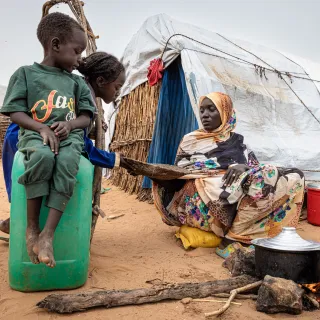 In a displacement camp in Sudan, a woman cooks over an open fire outside a makeshift shelter as two children watch.