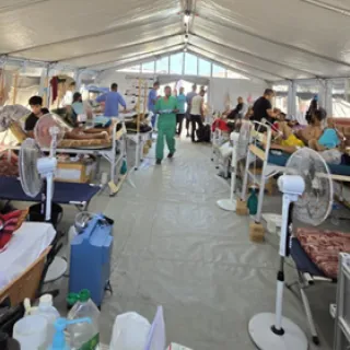 Patients lie on beds inside a crowded tent ward at the Red Cross Field Hospital in Rafah, Gaza. Medical staff in green scrubs move between beds, while fans are placed near patients to provide relief from the heat.