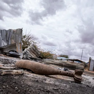 A rusted, unexploded mortar round lies on scorched ground in an abandoned area of Gedo’s Luuq district, Somalia. Behind it, twisted metal sheets and damaged structures sit under a heavy, overcast sky, highlighting the destruction left by recent fighting.