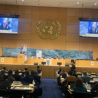 A large conference hall with delegates seated at long rows of desks facing a stage. Two large screens display a speaker at a podium, with the United Nations emblem prominently mounted on the wall behind him. Several panelists sit at a long table on the stage beneath a wide mural. Flags and conference banners are positioned to the side. The audience listens as a photographer captures the scene.
