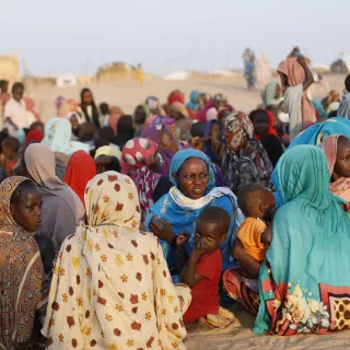 Crowd of women and children gathered outdoors in Tawila, North Darfur, many seated on the ground wearing colorful headscarves and traditional clothing, with a few people standing in the background against a dry, sandy landscape.