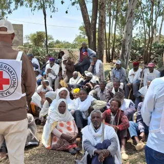 ICRC staff, seen from behind wearing Red Cross emblems, stand before a large seated crowd gathered outdoors.