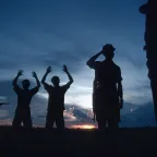 Two men standing with their hands raised up as another man points a weapon at them and two others look on