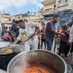Workers in white overalls stir large vats of food in the community kitchen, people and buildings in the background