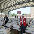 Two humanitarian workers (one from the ICRC and another one from the Yemeni Red Crescent) stand near each other surrounded by many piles of bags and boxes of food items