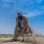 Churi Ibrahim, a 70-year-old farmer, works the dry, dusty soil with a hoe under a bright blue sky in a rural area, with modest shelters visible in the background — a scene reflecting resilience in the face of harsh living conditions.