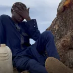 A young boy in a navy blue outfit sits at the base of a large tree, covering his face with one hand in a gesture of distress or exhaustion. A plastic water container rests beside him.