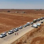 A long humanitarian aid convoy stretches along a rural road in Sweida, Syria, in July 2025. The convoy includes dozens of white vehicles marked with the Red Cross and Red Crescent emblems, along with large trucks carrying relief supplies. The road runs through a dry, reddish-brown landscape with sparse vegetation and scattered buildings in the distance. Aid workers in red uniforms walk near the vehicles under a clear blue sky.