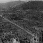 A black and white photograph showing the devastated landscape of Hiroshima after the nuclear bombing in August 1945. The area is flattened and burned, with only a few skeletal buildings and bare trees remaining. A straight dirt road runs through the center, surrounded by rubble and destruction, with mountains visible in the background.