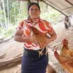 Mujer dentro de un gallinero, sostiene una gallina entre sus manos y sonríe a la cámara.