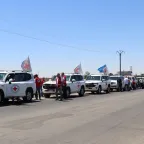A convoy of white vehicles marked with Red Cross emblems and flags parked on a road under a clear blue sky, with humanitarian workers standing beside them.