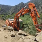 A man wearing a Red Crescent emblem points towards an excavator off a dirt road on a steep hillside in a mountainous area of Afghanistan, following the earthquake.