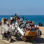 A crowded truck loaded with people and belongings drives along a coastal road, as others walk or travel on carts beside the sea.