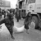 Two women carry a large sack of aid supplies together in front of Red Cross trucks, while other people unload and distribute relief items in the background