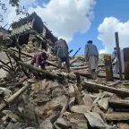 People search through the rubble of collapsed buildings after an earthquake, surrounded by broken wooden beams and stones, with mountains visible in the background under a partly cloudy sky.