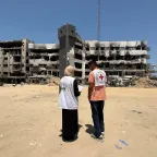 Two humanitarian workers wearing vests with Red Cross and partner organization logos stand in front of a heavily damaged, partially destroyed building, assessing the destruction in Gaza