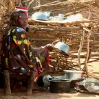 A woman washes dishes in an outdoor area surrounded by wooden fencing, with metal pots and a blue plastic bucket nearby.