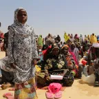 A group of people at an ICRC distribution in Niger. Most seated in the sand, one girl is standing in the foreground.